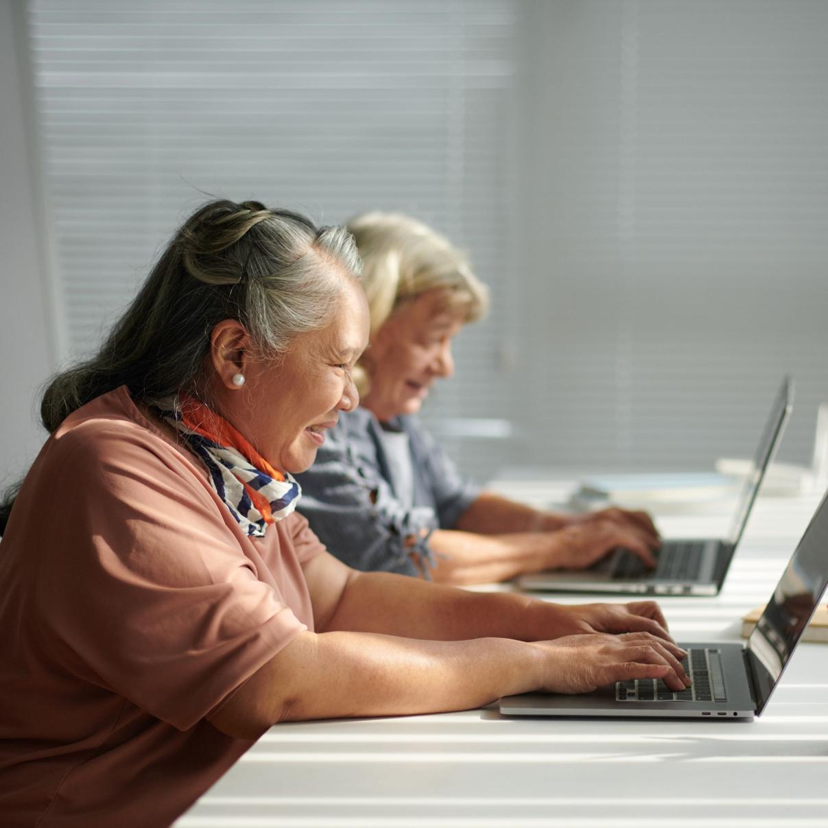 Two elderly women using laptops at a table with a blurred background