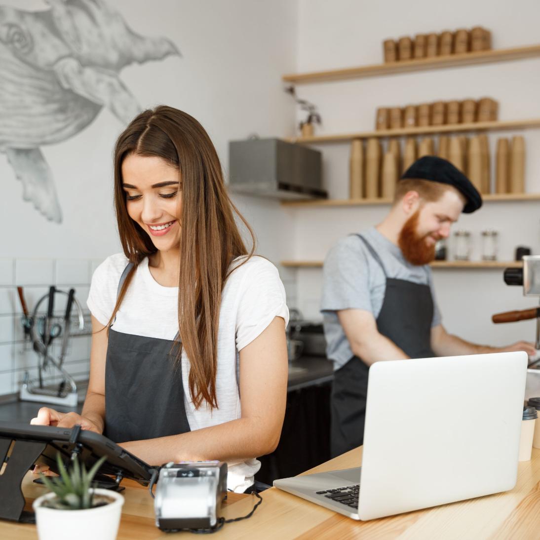 Two baristas working in a coffee shop with a laptop and cash register.